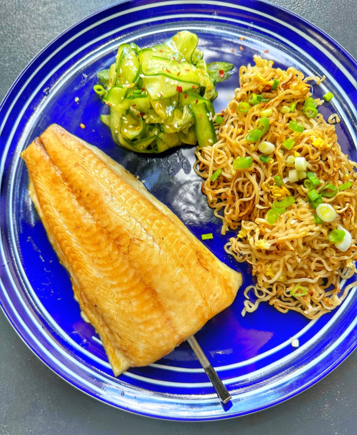 Black cod on a blue platter with stir fried noodles and cucumber salad.