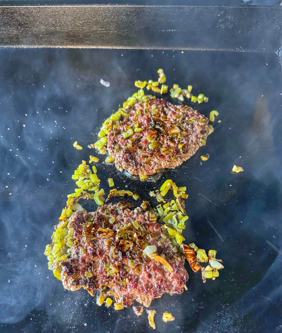 Two burger patties smashed with green chiles on the griddle.