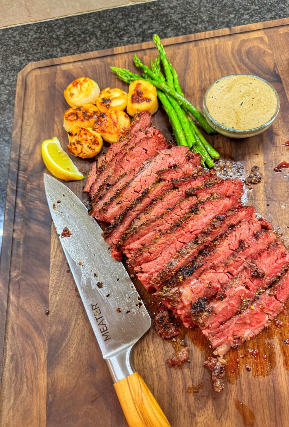 Reverse-seared Bavette steak sliced on a wooden cutting board, served with seared scallops, asparagus, and whiskey cream sauce.