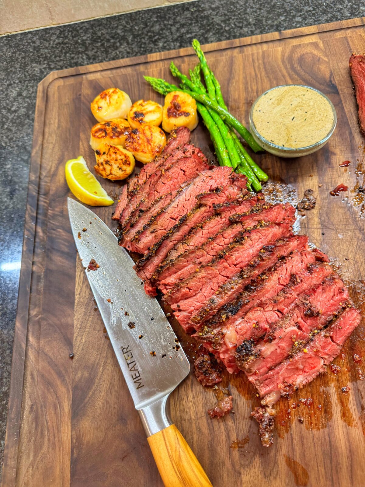 Reverse-seared Bavette steak sliced on a wooden cutting board, served with seared scallops, asparagus, and whiskey cream sauce.