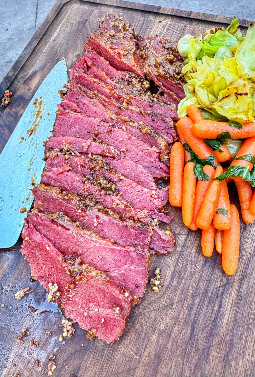 Sliced maple mustard corned beef on a wooden cutting board with braised cabbage and maple basil carrots.