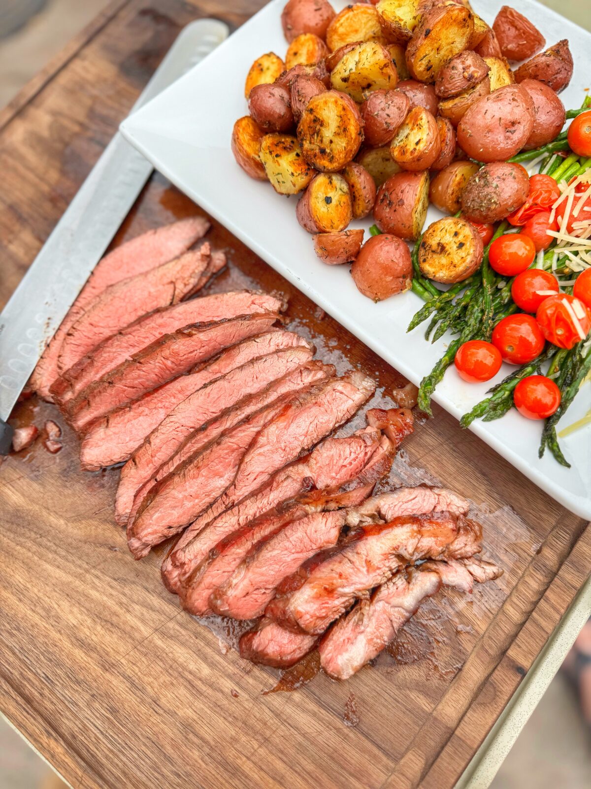 Sliced grilled flank steak on a cutting board next to a plate of roasted potatoes and vegetables.