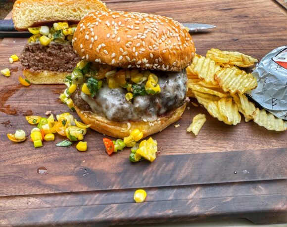 Finished butter burger on sesame bun with corn slaw and chips on wooden board.