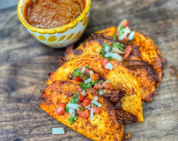 Three crispy birria tacos on a wooden board with a bowl of consommé.
