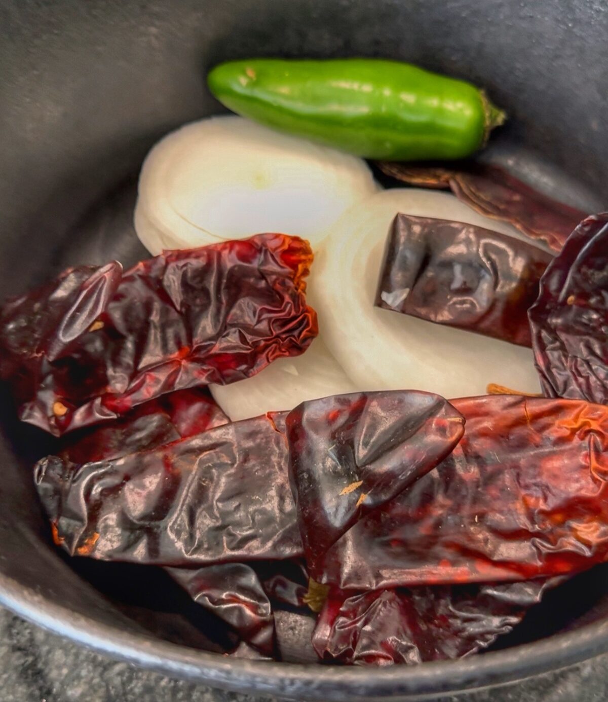 Dried guajillo chiles, sliced white onion, and a green jalapeño in a black pan.