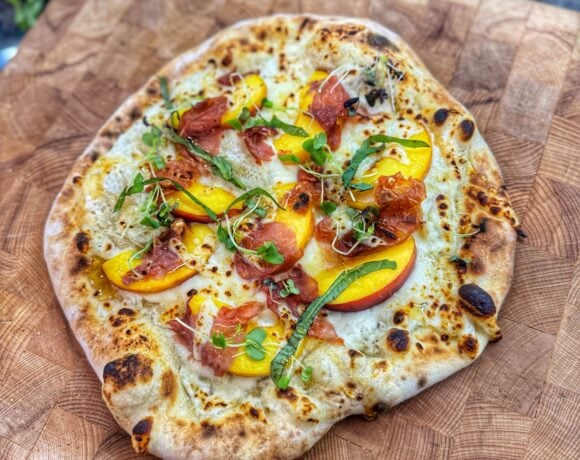 Golden brown pizza topped with peaches, prosciutto, melted cheese, and microgreens.