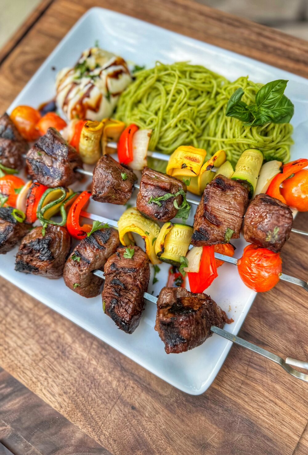 Plated herb-marinated steak and veggie skewers served with pesto angel hair pasta and burrata.