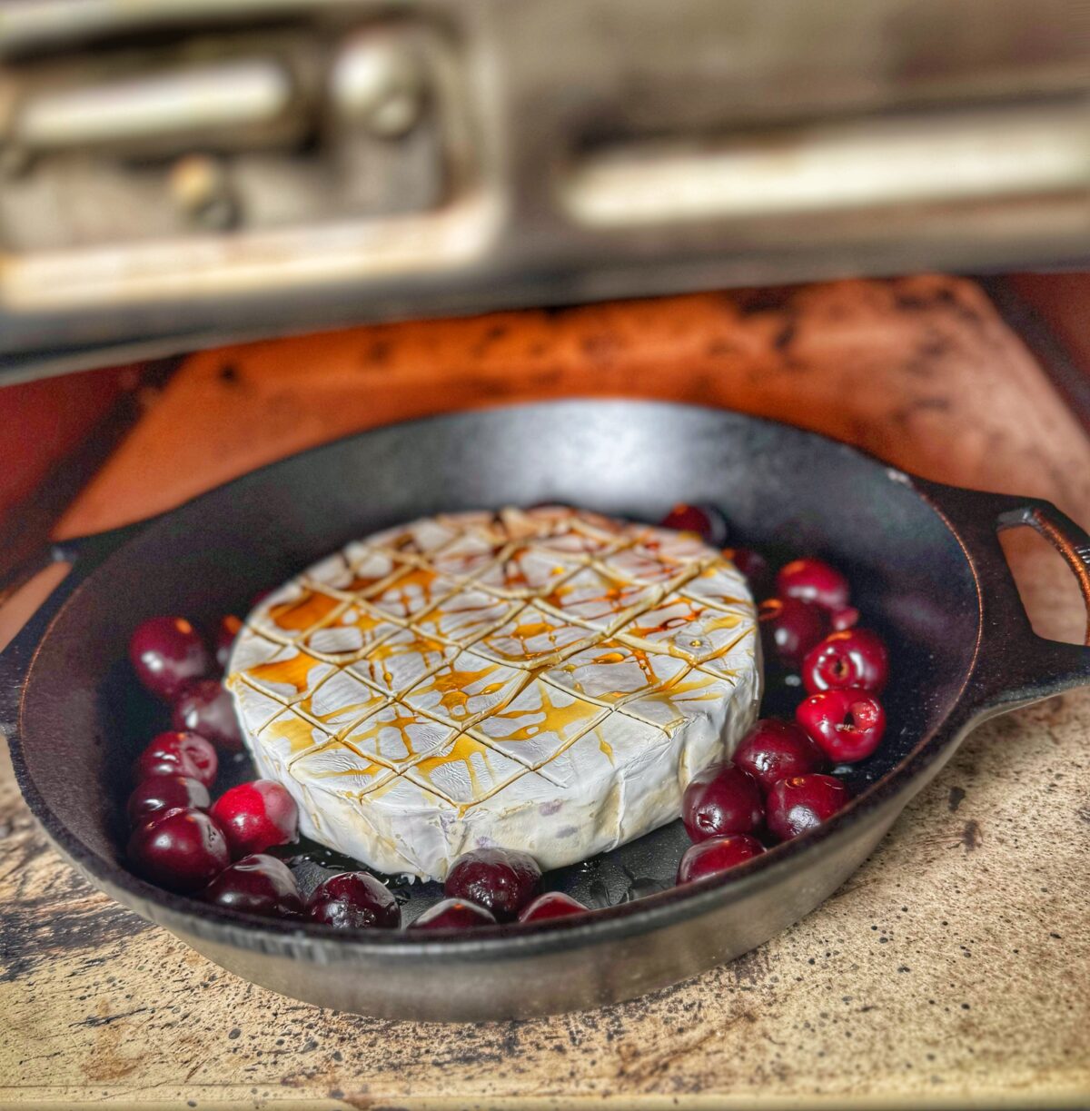 Brie scored on top with cherries around it in a cast iron skillet, baking inside the Ooni Karu Pro oven.