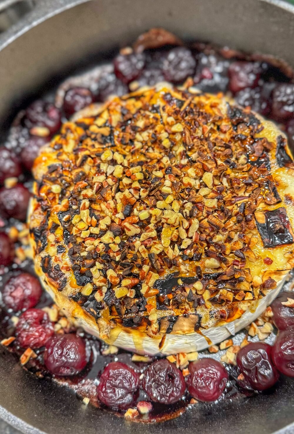Baked brie topped with chopped pecans in a cast iron skillet surrounded by cherries.