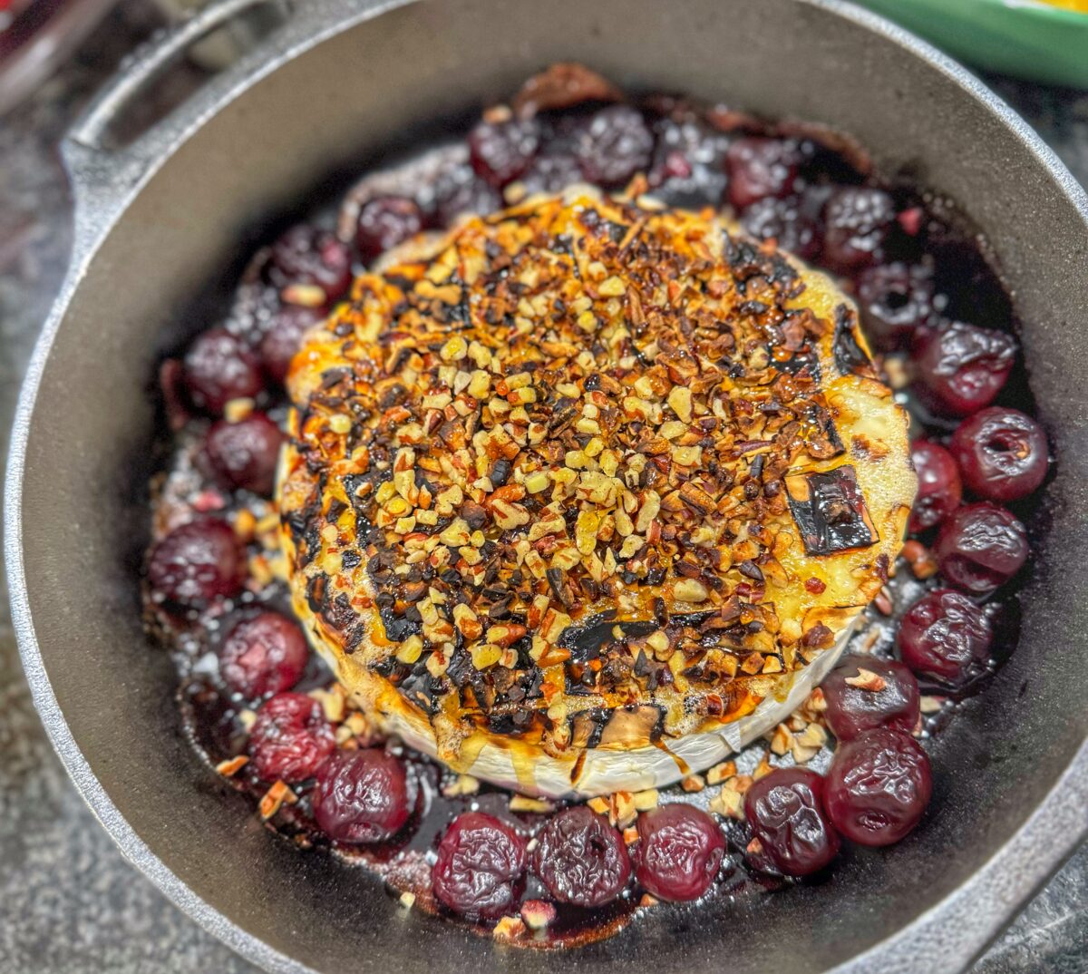 Baked brie topped with chopped pecans in a cast iron skillet surrounded by cherries.