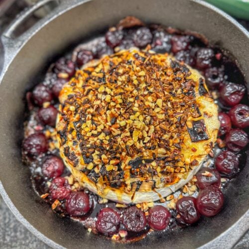 Baked brie topped with chopped pecans in a cast iron skillet surrounded by cherries.