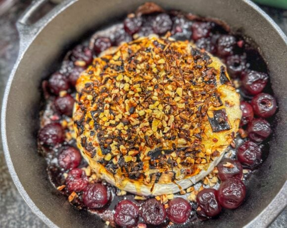 Baked brie topped with chopped pecans in a cast iron skillet surrounded by cherries.