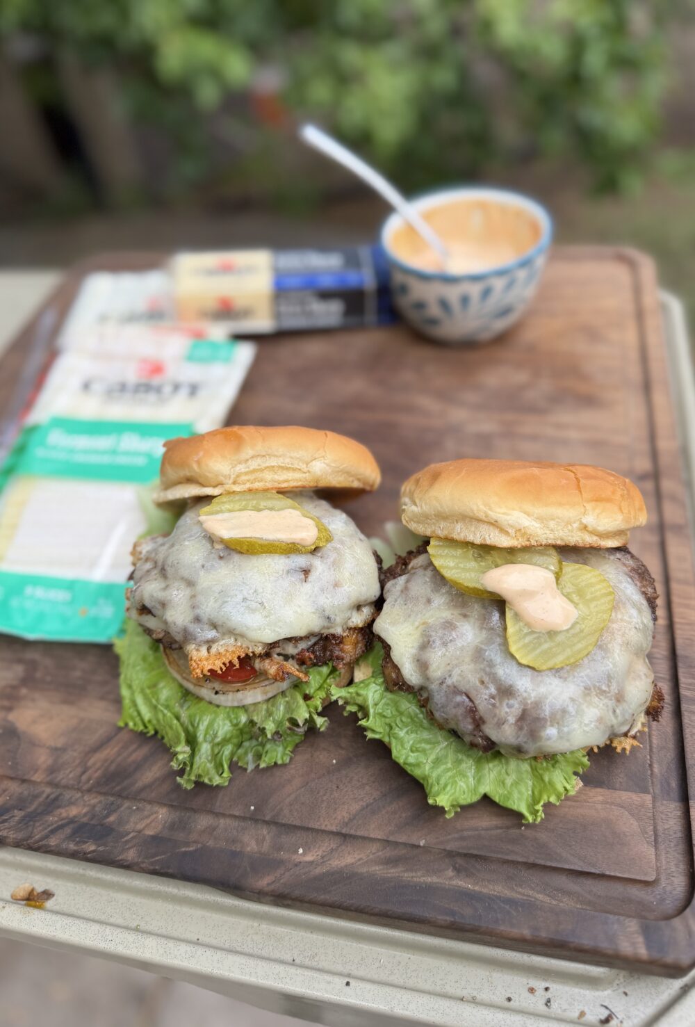 Two smashburgers on a cutting board topped with melted cheddar, pickles, and burger sauce, with Cabot cheddar packages and sauce bowl in the background.
