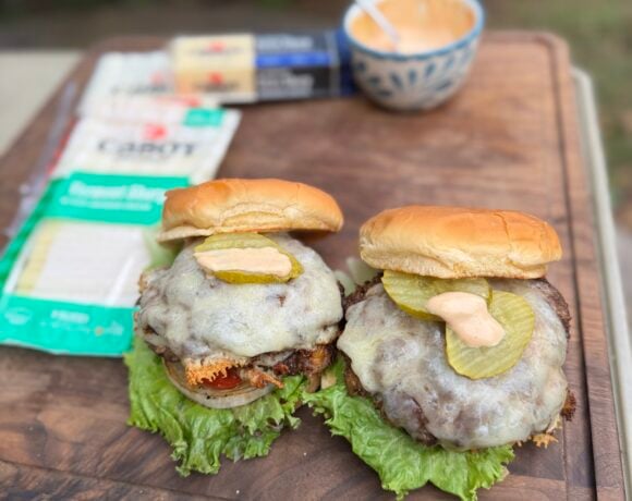 Two smashburgers on a cutting board topped with melted cheddar, pickles, and burger sauce, with Cabot cheddar packages and sauce bowl in the background.