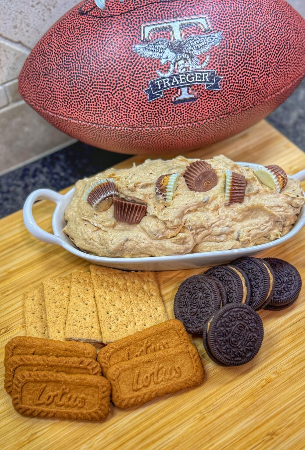 Dish of peanut butter cup cheesecake dip with peanut butter cups on top, surrounded by Oreos, graham crackers, and Lotus Biscoff cookies, next to a Traeger football.