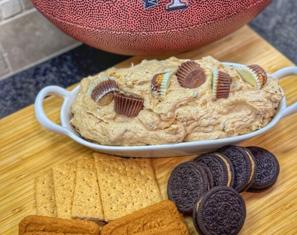 Dish of peanut butter cup cheesecake dip with peanut butter cups on top, surrounded by Oreos, graham crackers, and Lotus Biscoff cookies, next to a Traeger football.