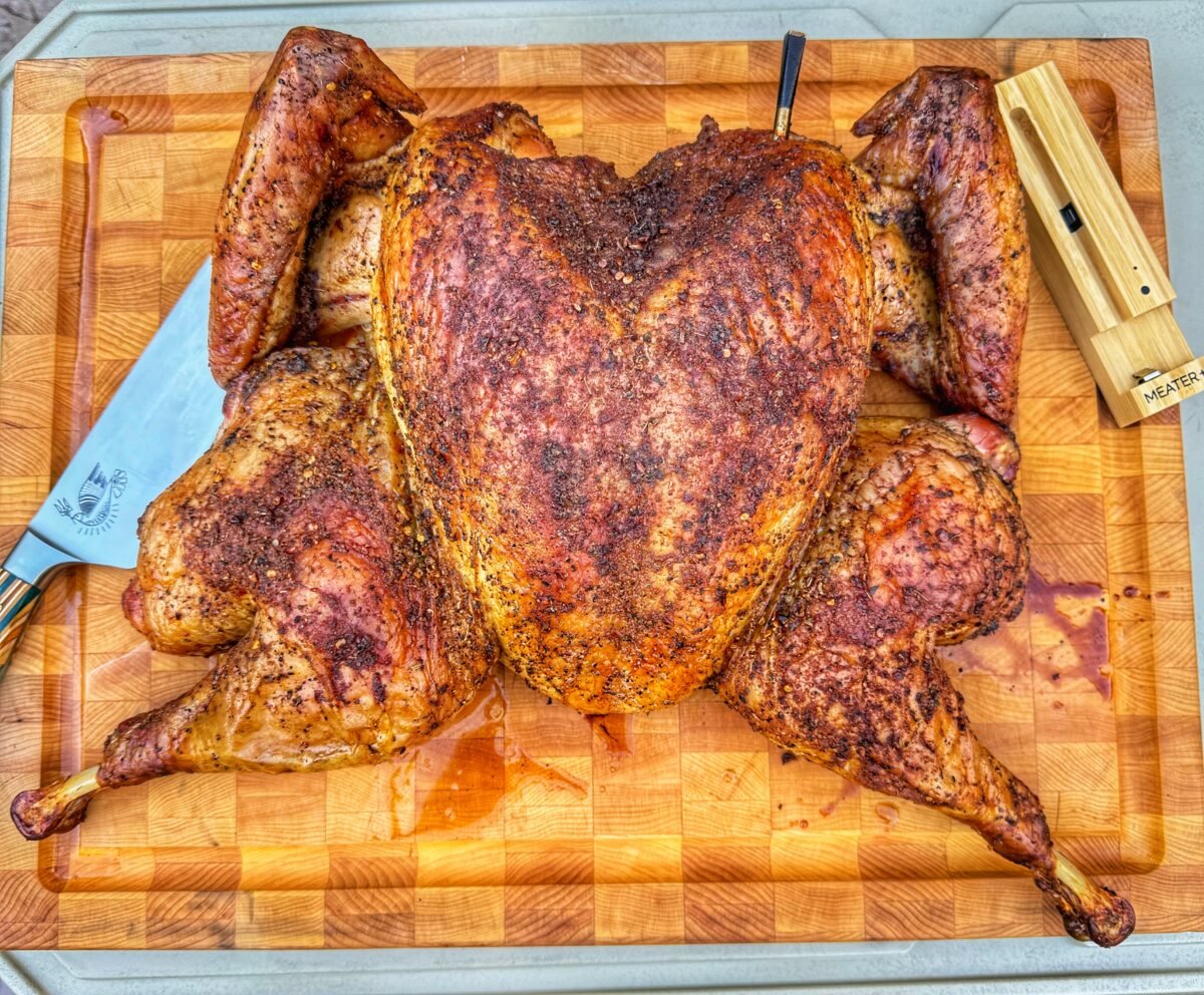 Fully cooked spatchcock turkey resting on a wooden cutting board with carving knife and thermometer.