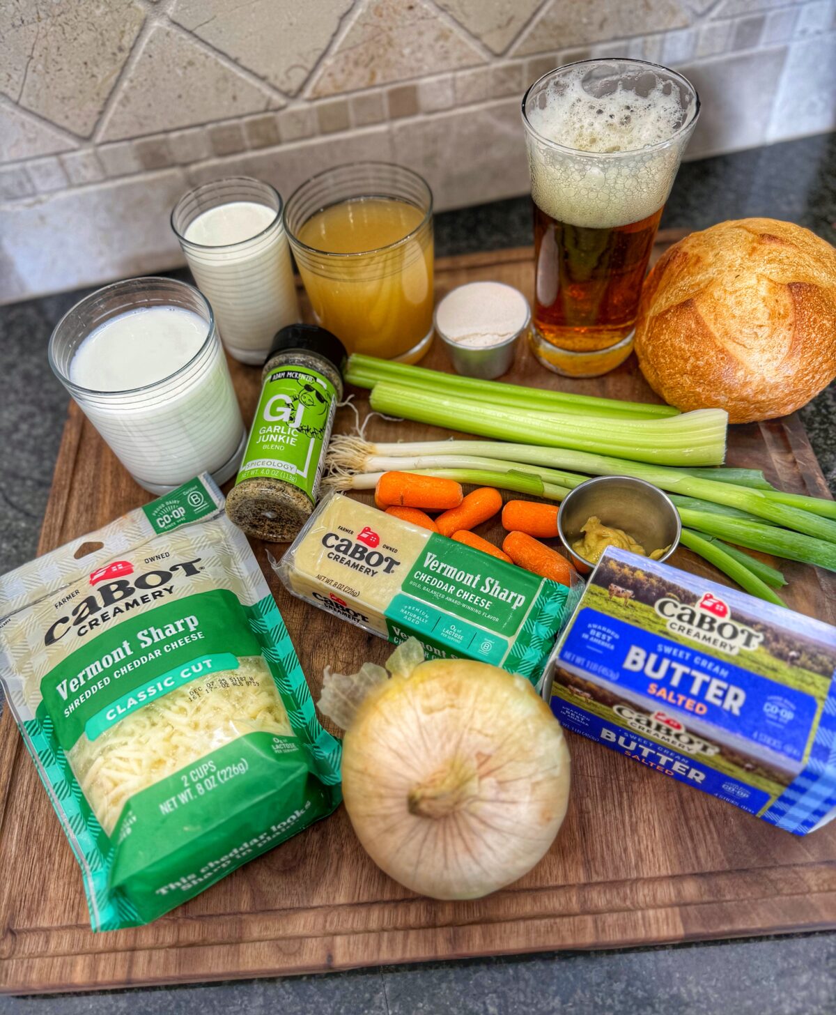 Ingredients for beer cheese soup on a wooden board, including Cabot Creamery cheddar, butter, onion, celery, carrots, milk, broth, beer, and spices.