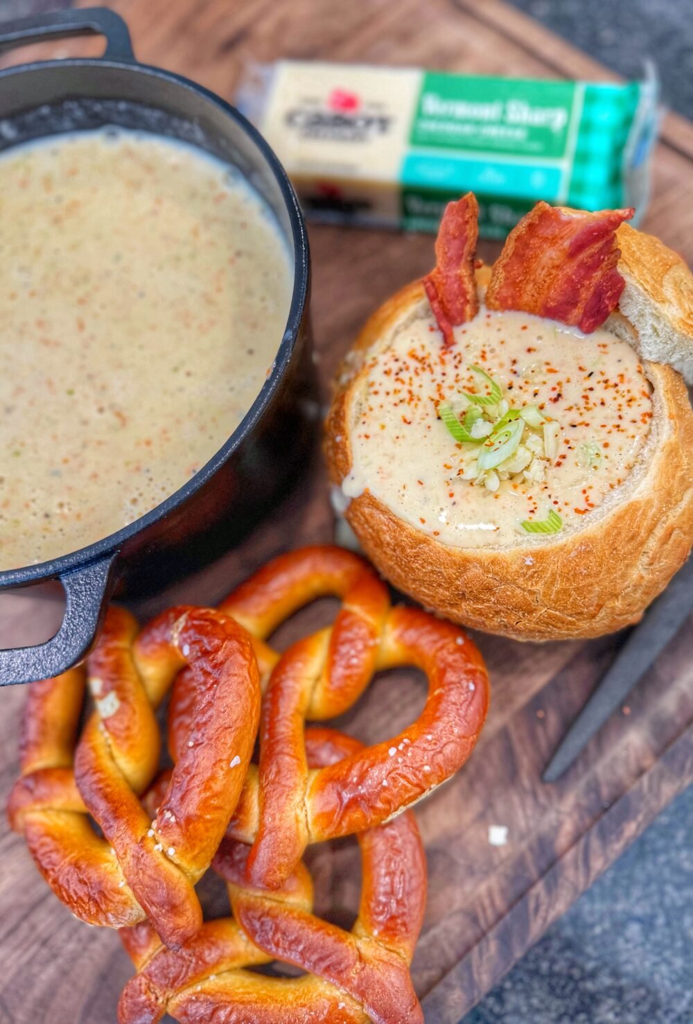 A close-up bread bowl of creamy soup topped with shredded cheese, bacon pieces, and chopped green onions.