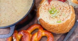 A close-up bread bowl of creamy soup topped with shredded cheese, bacon pieces, and chopped green onions.