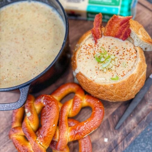 A close-up bread bowl of creamy soup topped with shredded cheese, bacon pieces, and chopped green onions.