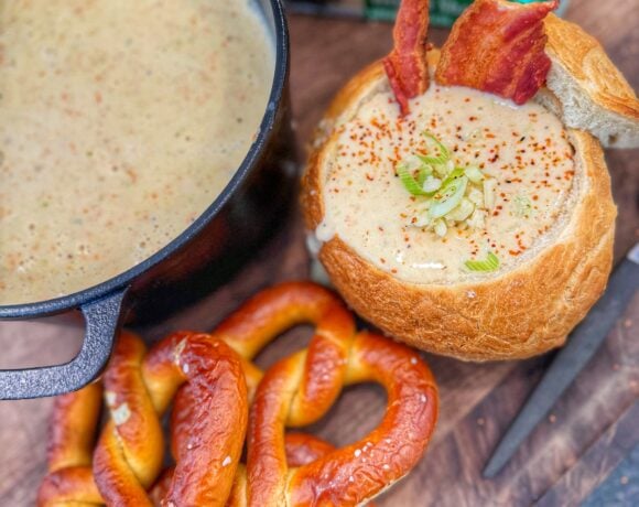 A close-up bread bowl of creamy soup topped with shredded cheese, bacon pieces, and chopped green onions.