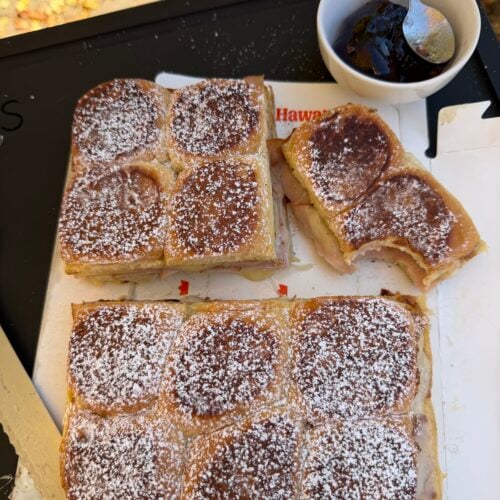 Monte Cristo sliders cut into sections on a tray, dusted with powdered sugar, served with grape jelly in a bowl.
