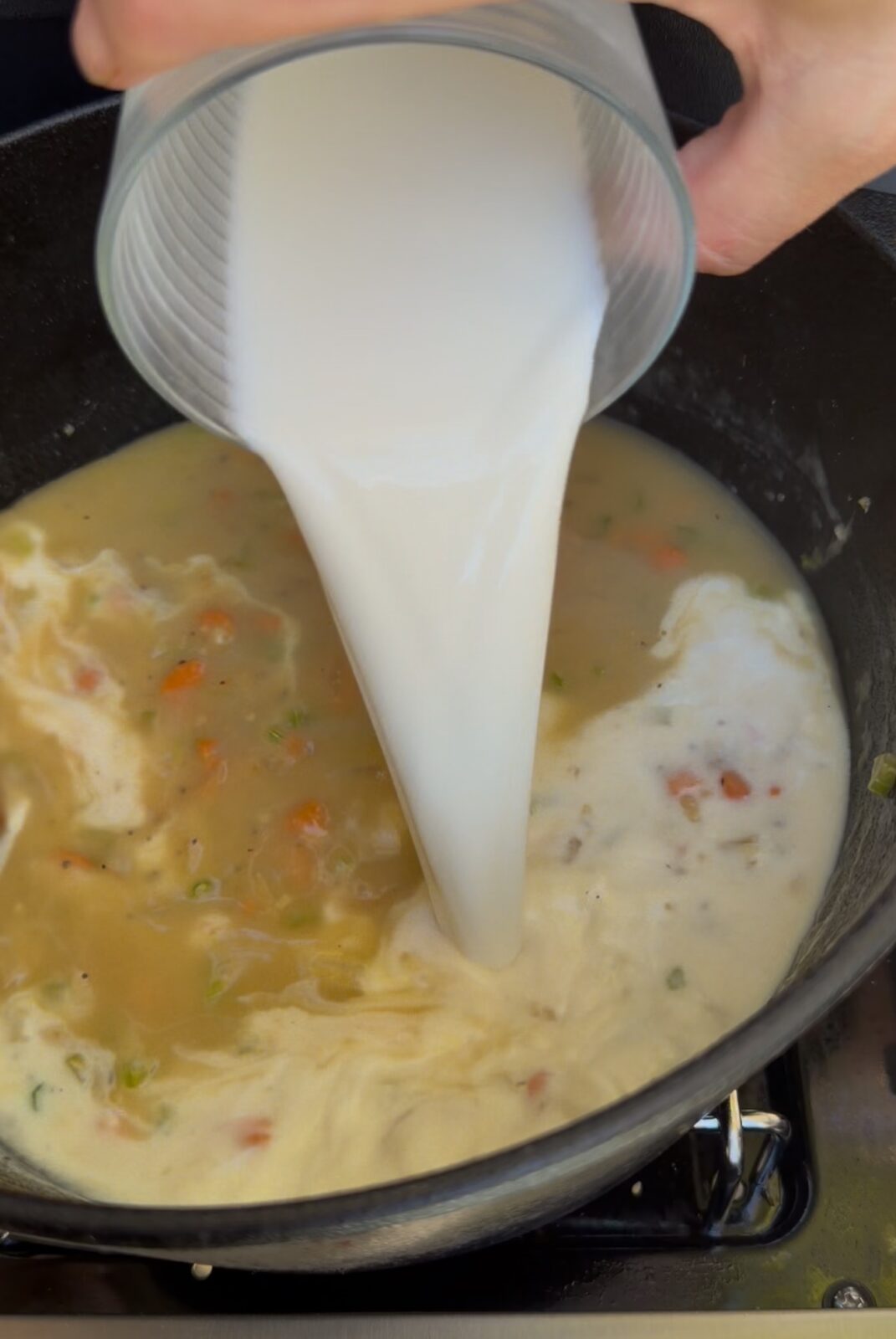 A glass of milk being poured into a pot of soup with vegetables.
