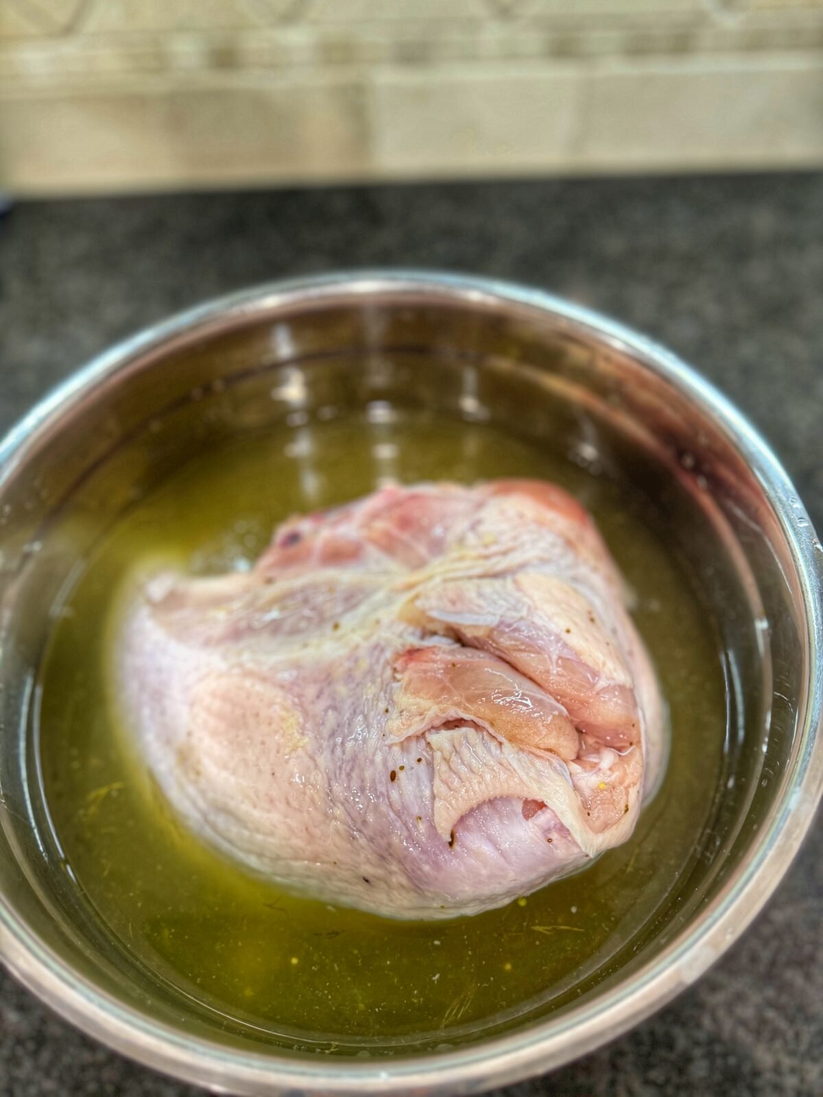 A bone-in turkey breast fully submerged in greenish pickle brine inside a stainless steel mixing bowl.