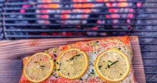 Salmon fillet topped with lemon slices and herbs on a cedar plank inside a charcoal grill over glowing coals.