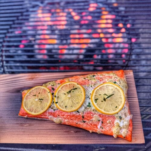 Salmon fillet topped with lemon slices and herbs on a cedar plank inside a charcoal grill over glowing coals.