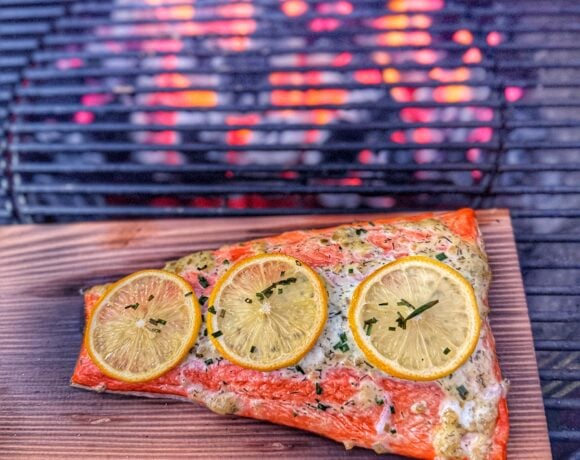 Salmon fillet topped with lemon slices and herbs on a cedar plank inside a charcoal grill over glowing coals.
