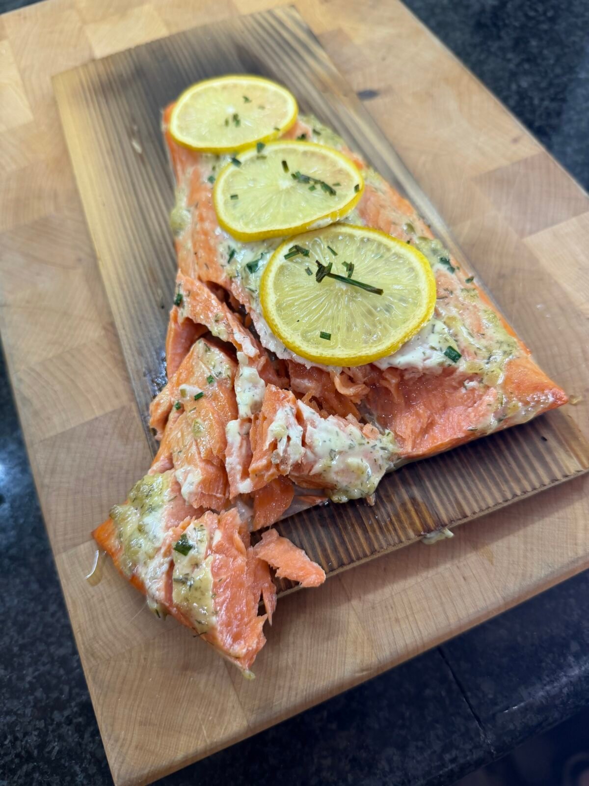 Cooked cedar plank salmon topped with lemon slices and herbs, with pieces flaked off onto the wooden board.