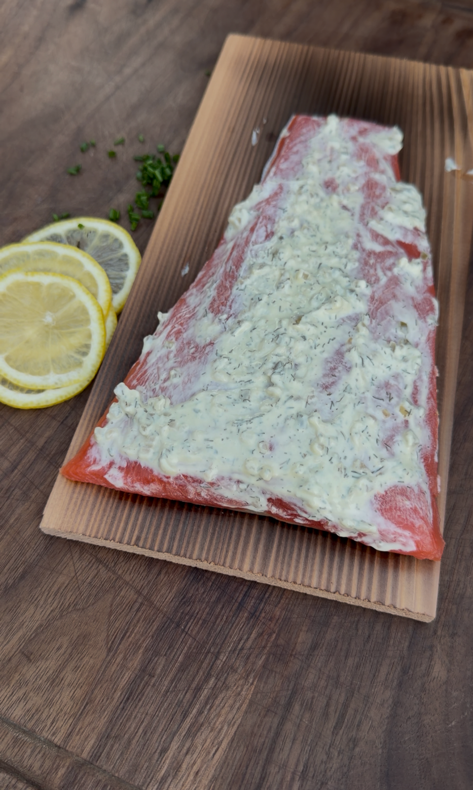 Uncooked salmon fillet on a cedar plank coated with a creamy dill sauce, with sliced lemons and chopped chives on the wooden surface beside it.