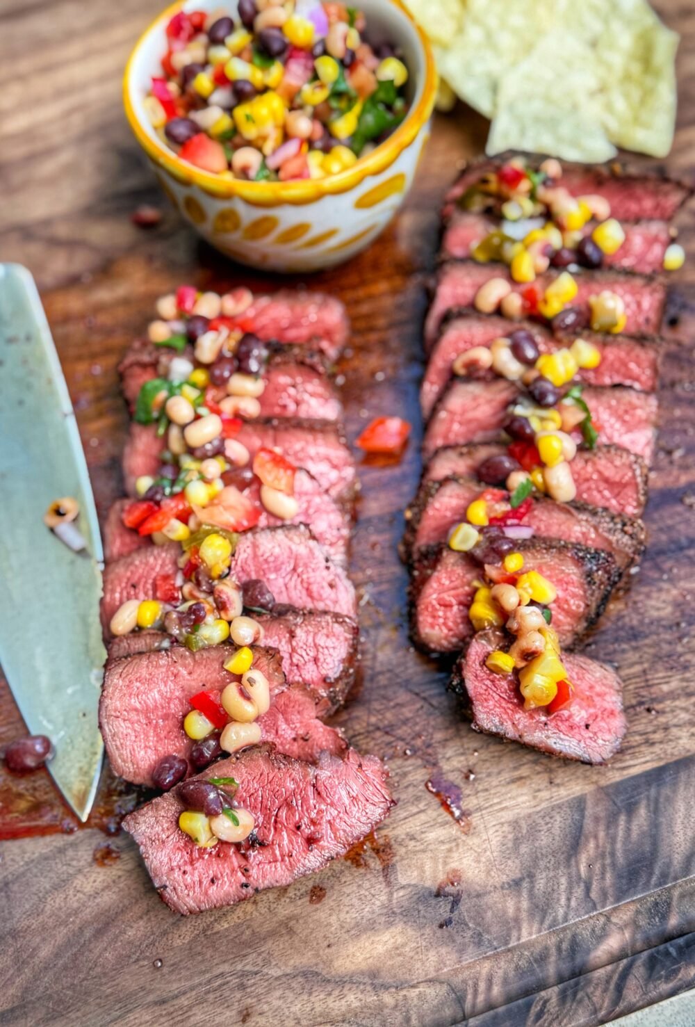 Sliced steak topped with cowboy caviar on a wooden cutting board with tortilla chips and a bowl in the background