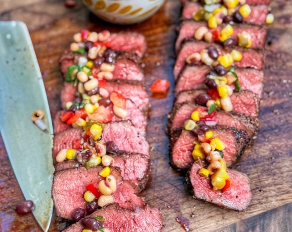 Sliced steak topped with cowboy caviar on a wooden cutting board with tortilla chips and a bowl in the background