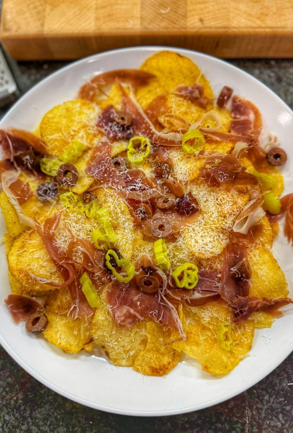 Charcuterie nachos made with kettle potato chips topped with prosciutto, finely grated cheese, sliced pepperoncini, and olives on a white plate