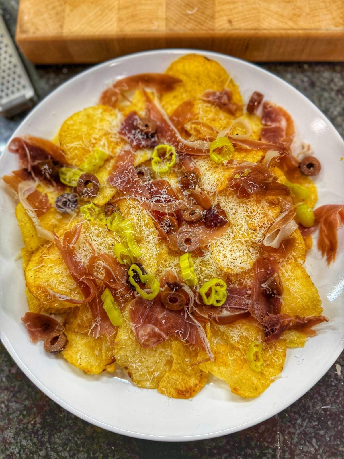 Charcuterie nachos made with kettle potato chips topped with prosciutto, finely grated cheese, sliced pepperoncini, and olives on a white plate