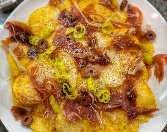 Charcuterie nachos made with kettle potato chips topped with prosciutto, finely grated cheese, sliced pepperoncini, and olives on a white plate