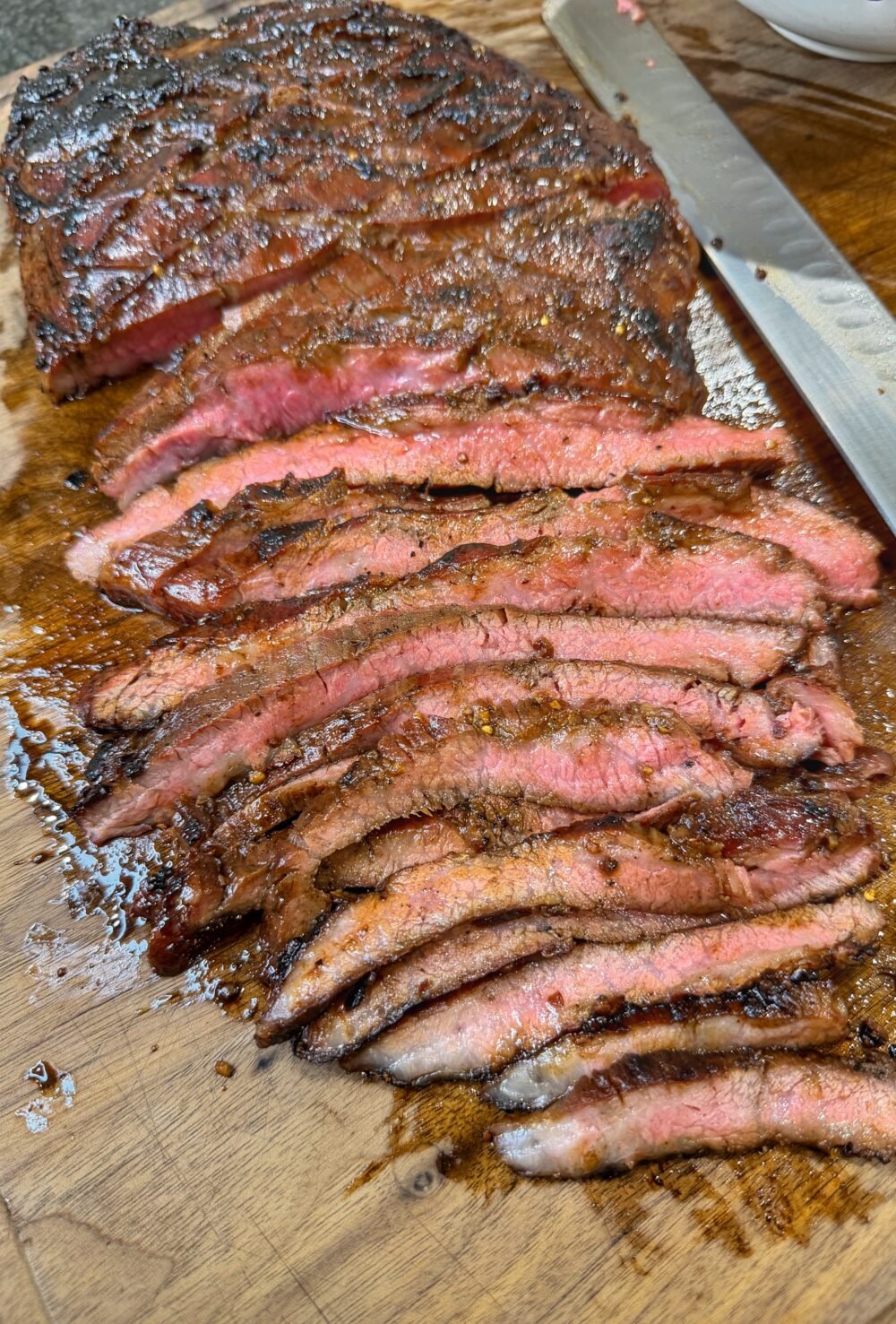 Sliced cooked flank steak arranged on a wooden cutting board with a knife nearby.