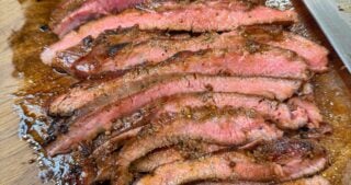 Sliced cooked flank steak arranged on a wooden cutting board with a knife nearby.