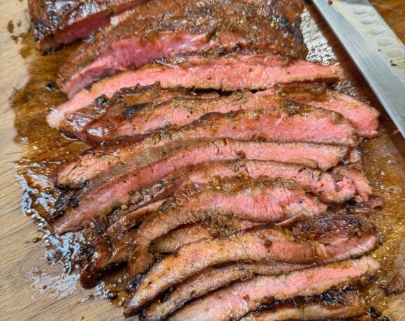 Sliced cooked flank steak arranged on a wooden cutting board with a knife nearby.