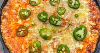 Cast iron skillet filled with chili cheese hot dog dip topped with sliced jalapeños, melted cheese, and visible chili oil on the surface.