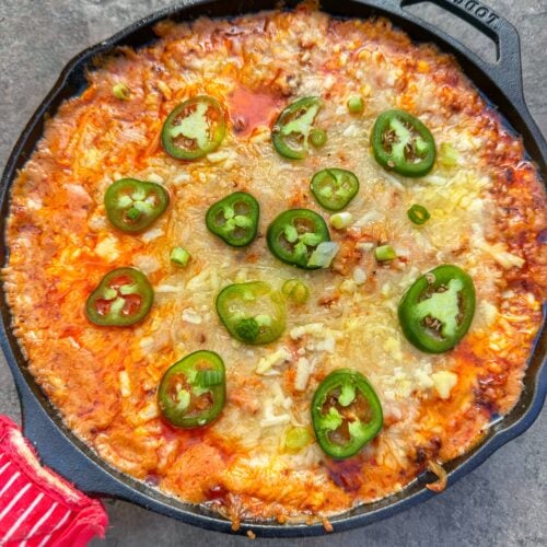Cast iron skillet filled with chili cheese hot dog dip topped with sliced jalapeños, melted cheese, and visible chili oil on the surface.