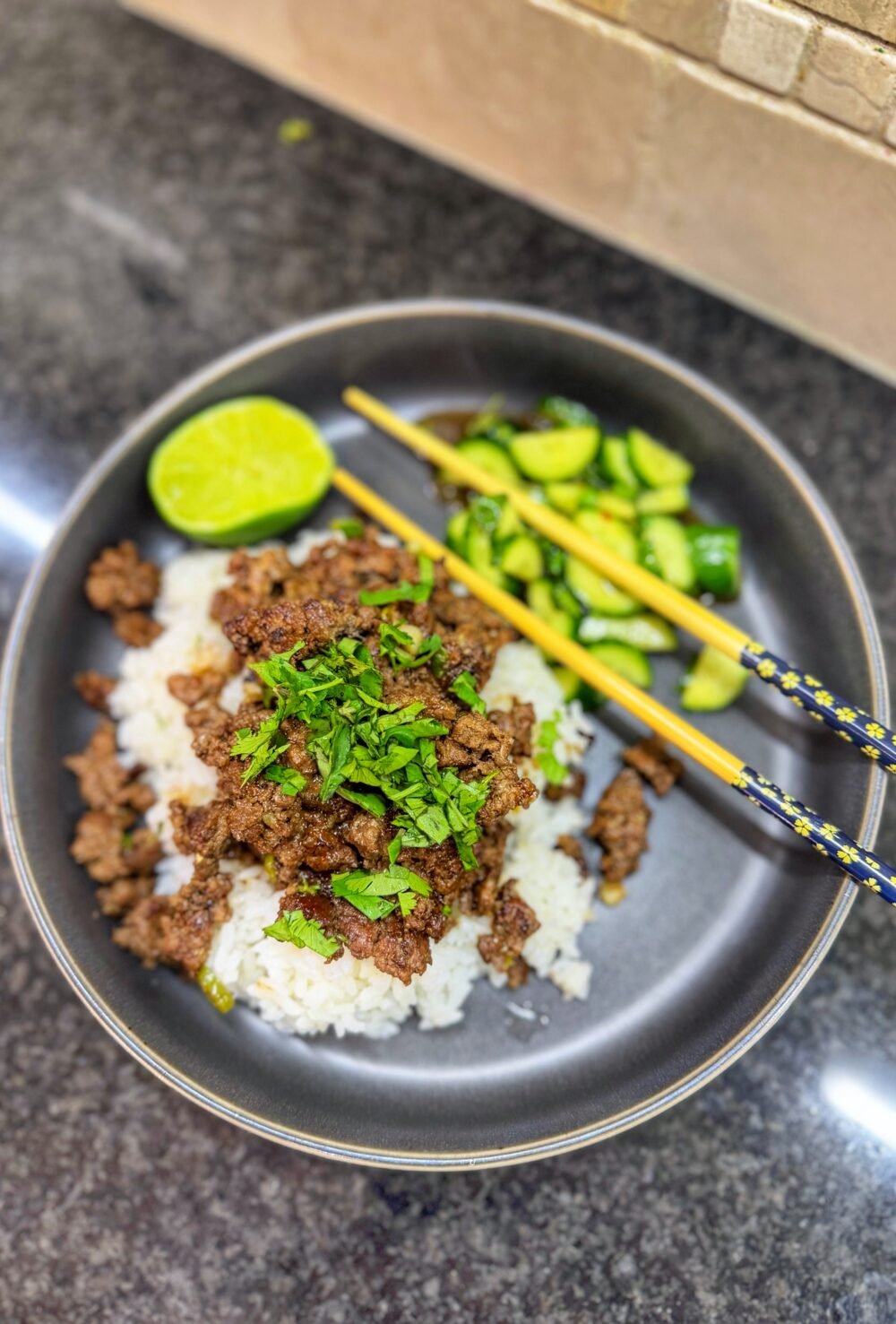 Bowl of white rice topped with ground beef and cilantro served with cucumber salad and lime.