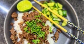 Bowl of white rice topped with ground beef and cilantro served with cucumber salad and lime.