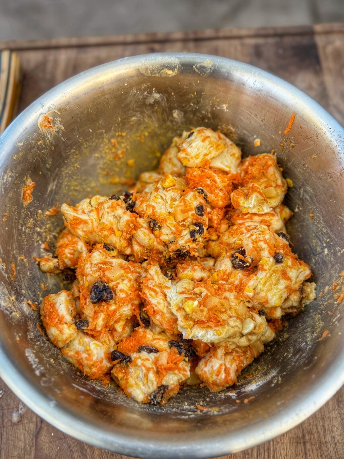 Biscuit dough pieces mixed with shredded carrots, raisins, pineapple, and brown sugar in a large metal bowl.