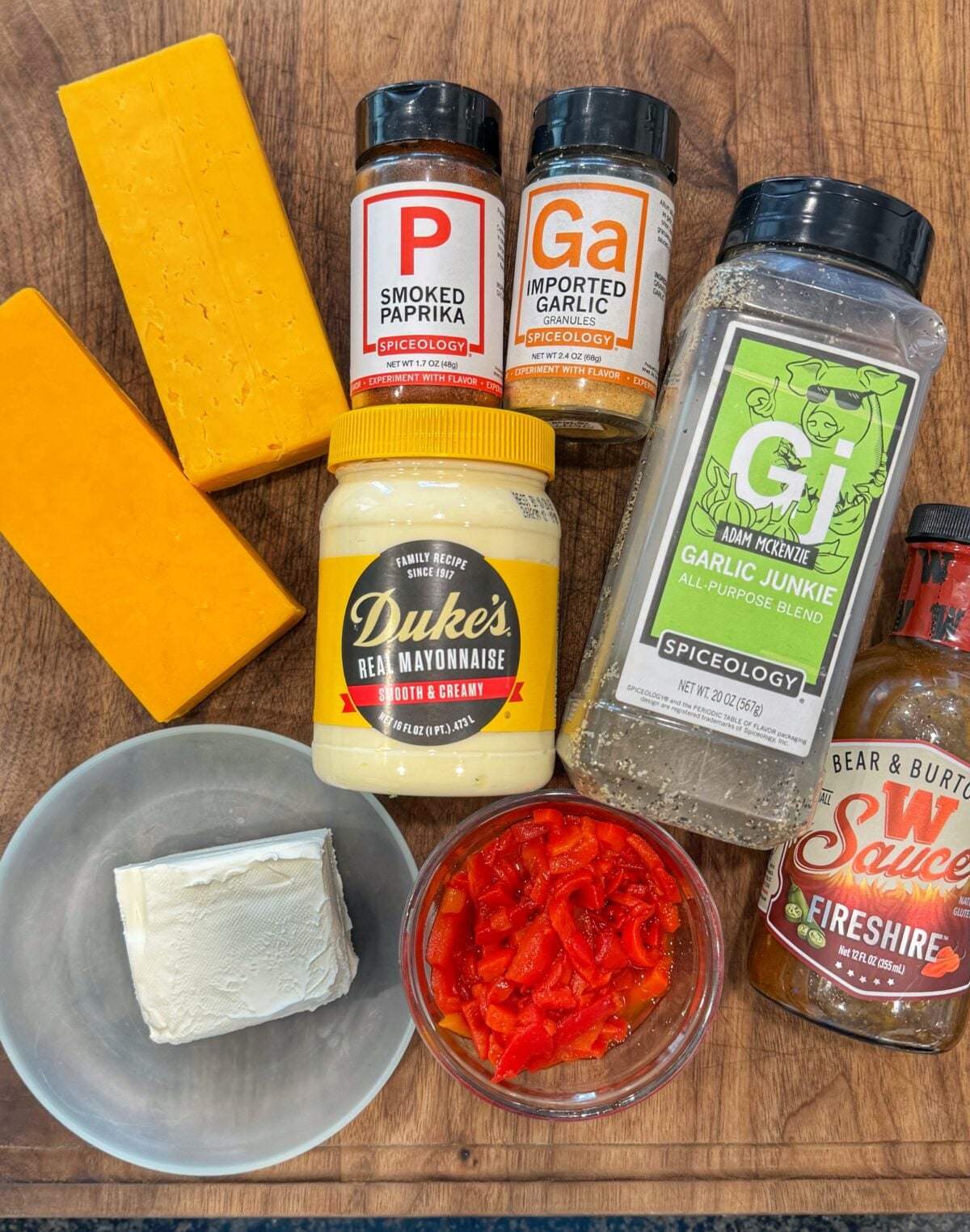 Blocks of cheddar cheese, Duke’s mayonnaise, cream cheese, diced pimentos, smoked paprika, garlic seasoning, garlic blend seasoning, and Worcestershire sauce arranged on a wooden board.