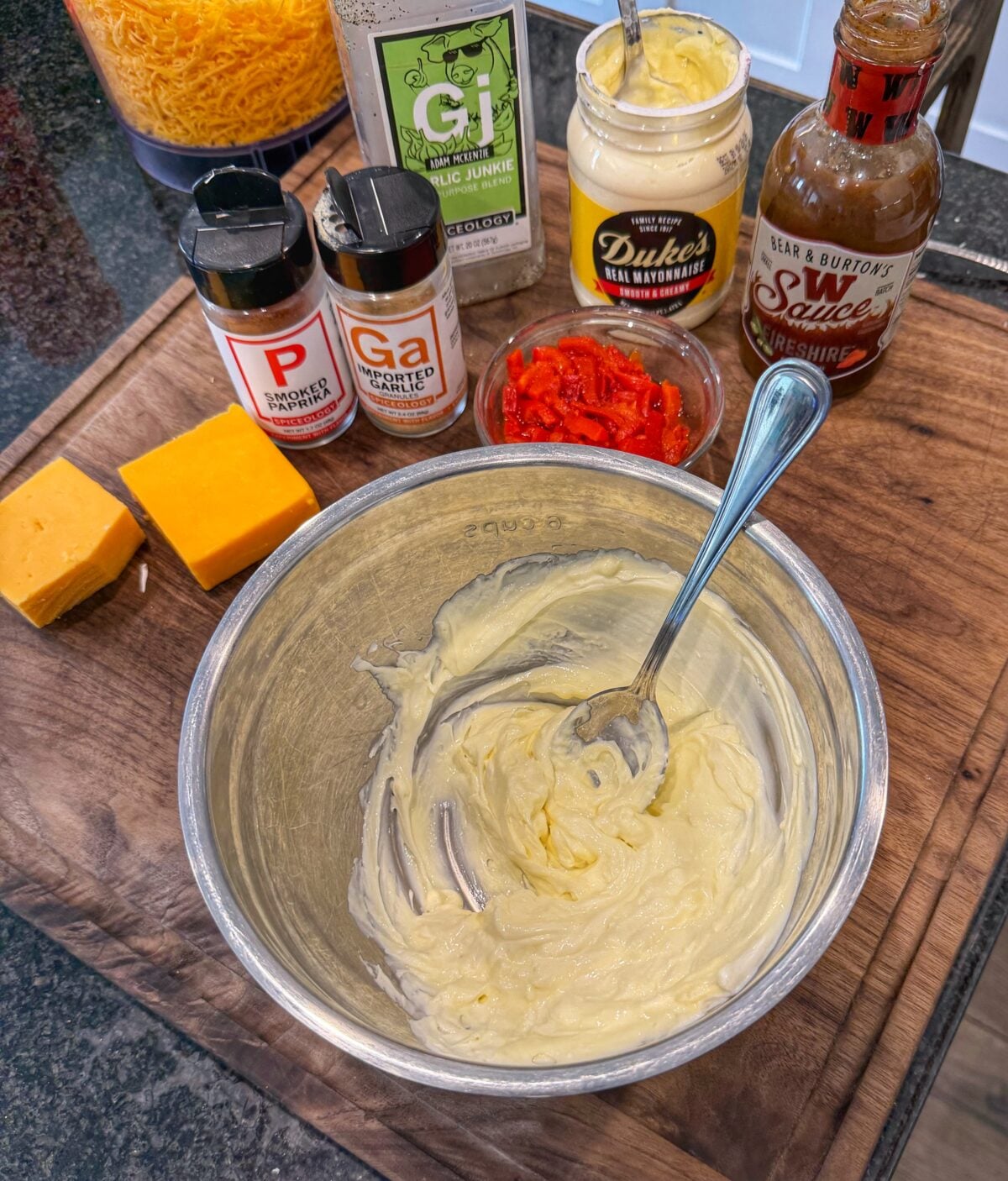 Cream cheese and mayonnaise mixed together in a metal bowl with shredded cheddar cheese, diced pimentos, and seasonings visible in the background.