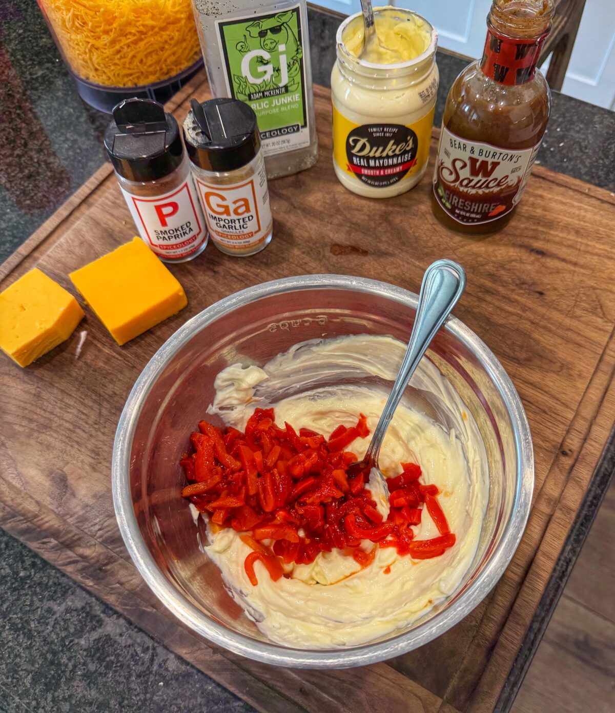Diced pimentos added to a creamy mixture of cream cheese and mayonnaise in a metal mixing bowl.